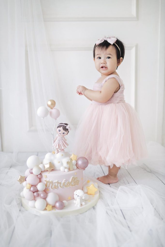 Baby girl standing in a soft pink dress with a bright white studio background.