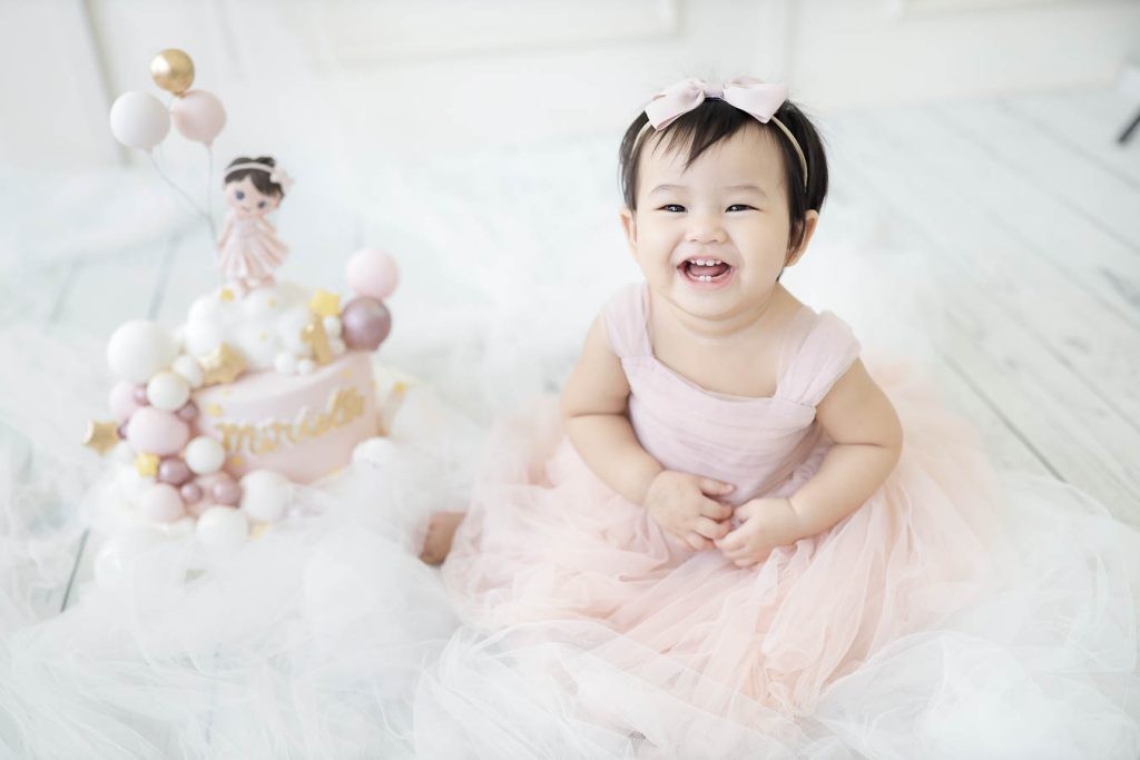 Baby girl wearing a pastel pink dress sitting on soft white fabric in a minimalist studio setting