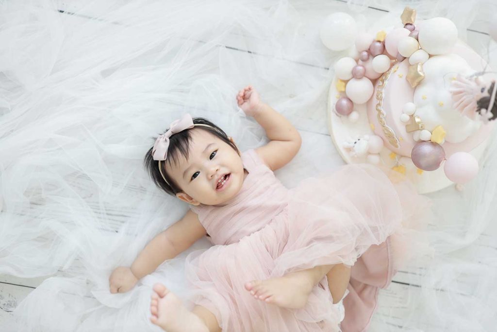 Baby girl lying down and smiling, wearing a pink pastel dress with a matching headband